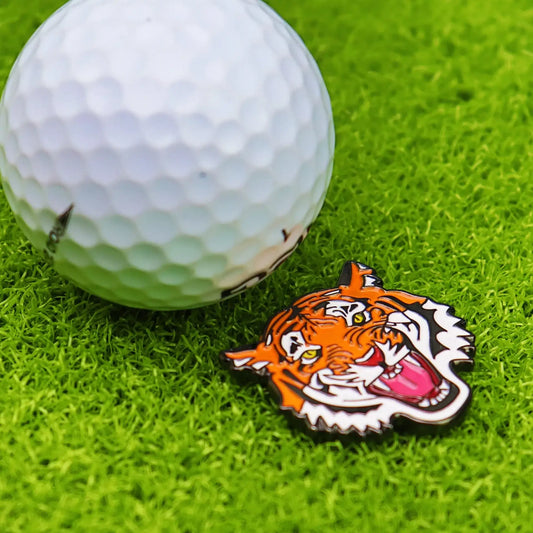 Golf ball marker hat clip featuring a tiger design beside a white golf ball on green grass.